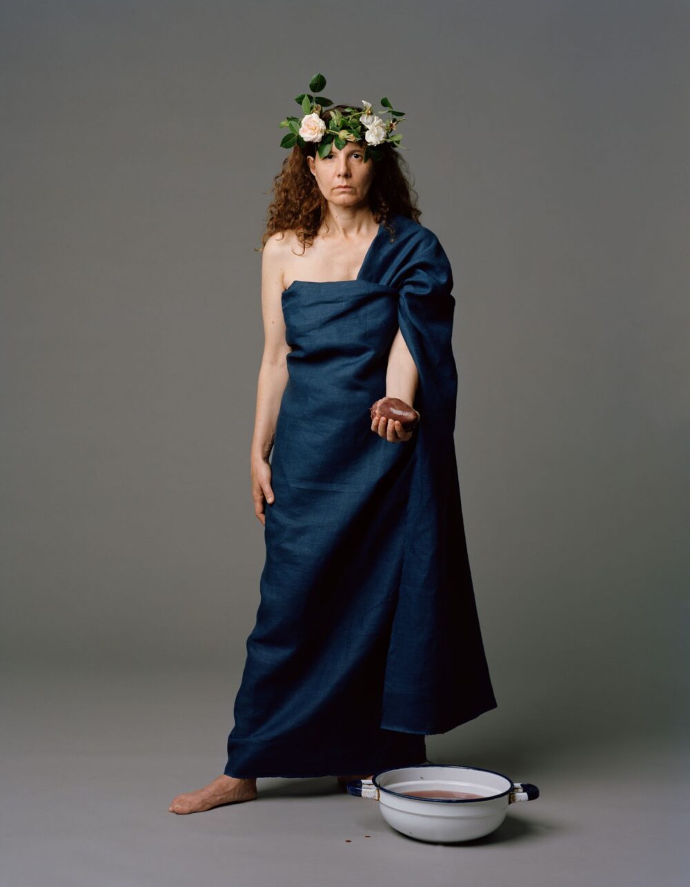 Foto einer Frau, in ein blaues Tuch gehüllt, mit einer Blumenkrone auf dem Kopf und einem blutigen Herz in der Hand.