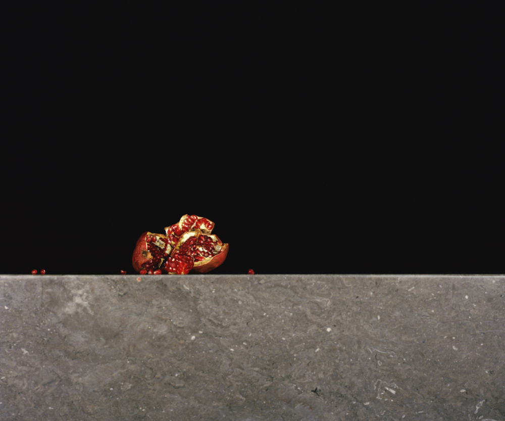 Fotografie: Ein aufgebrochener Granatapfel liegt vor schwarzem Hintergrund auf einr grauen Steinmauer, einzelne Granatapfelgerne liegen links und rechts verteilt.