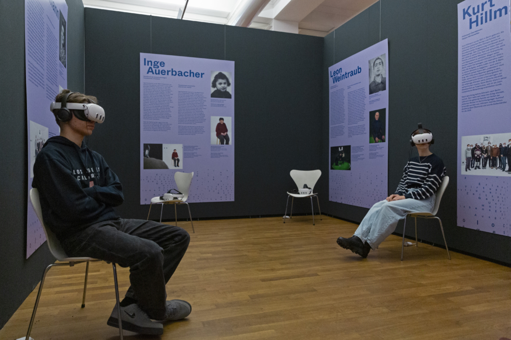 Zwei junge Besucher*innen sitzen mit VR-Brillen auf Stühlen in der Ausstellung KZ überlebt.